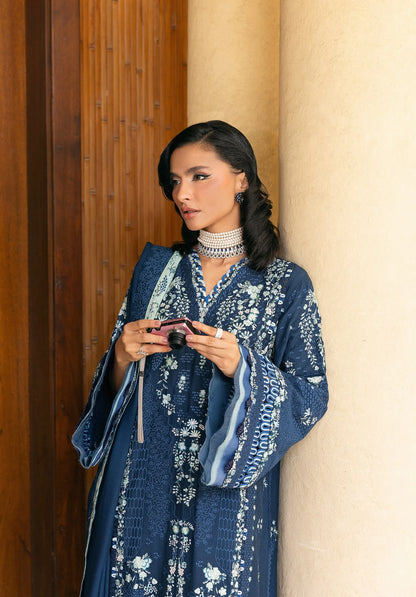 Woman in a blue embroidered outfit standing against a beige wall.