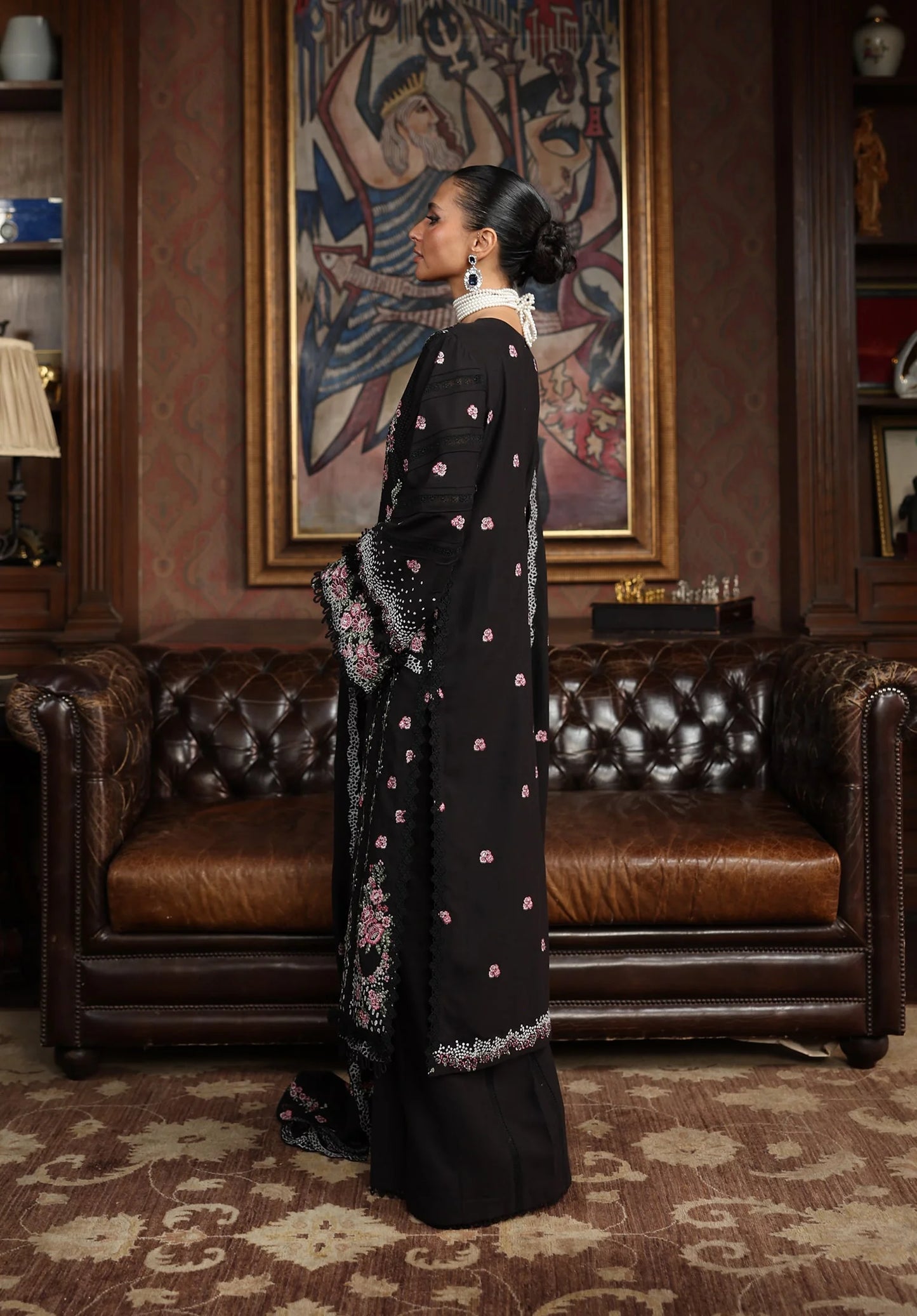 Woman in a black dress with floral patterns standing in an ornate room with a brown leather couch and decorative wall art.