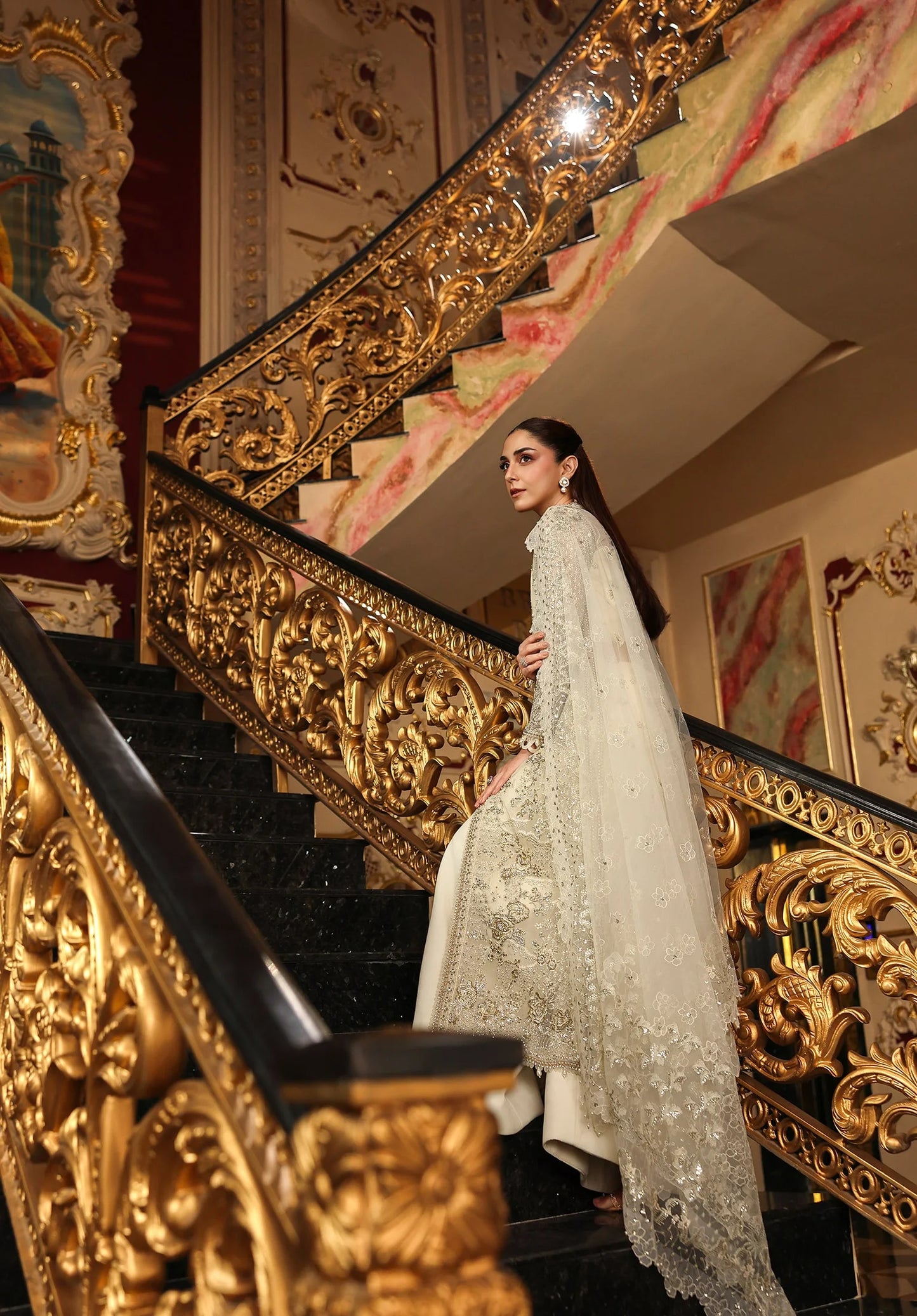 Woman in a white embroidered outfit standing on a grand staircase with gold railings.