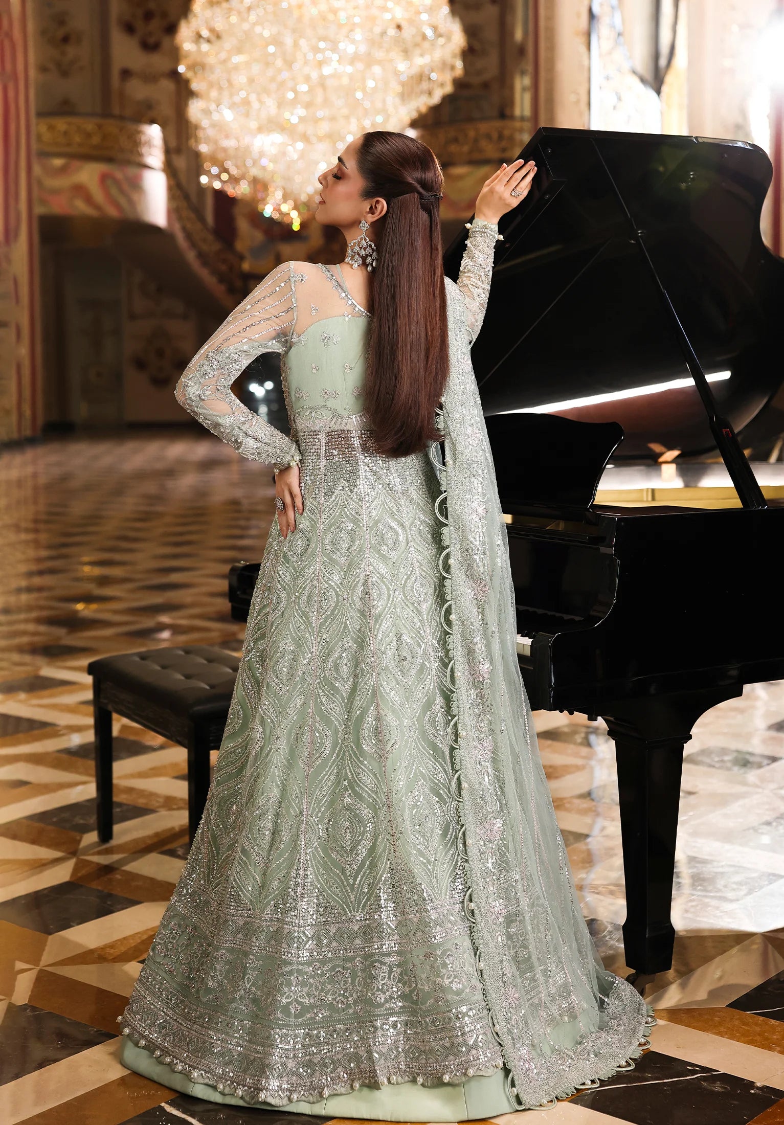 Woman in a light green embroidered dress standing next to a grand piano in an elegant room with Elaf Stardom Handwork.