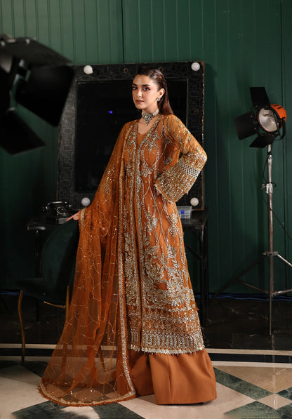 Woman in an orange embroidered traditional outfit standing in a studio setting with green walls and lighting equipment with Elaf Stardom Handwork